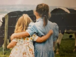 A picture of two children with their arms wrapped around each other and their backs to the camera, looking at a field of cows